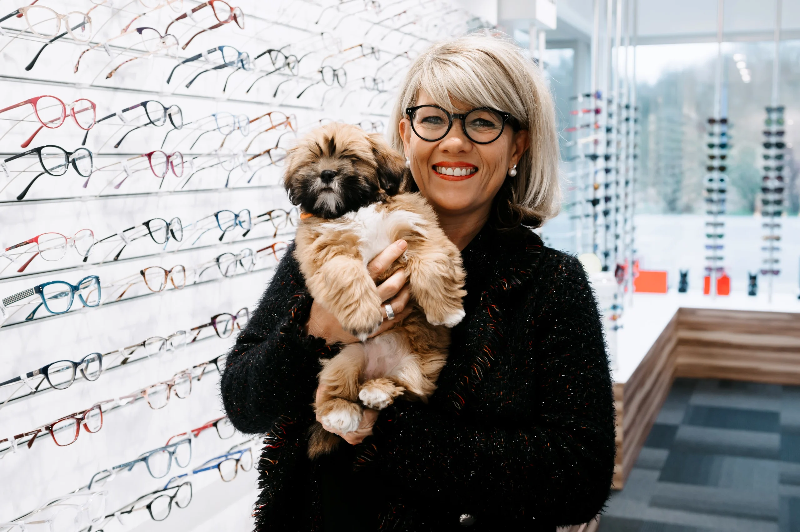 Femme élégante et souriante portant un petit chien devant un présentoir de lunettes de vue