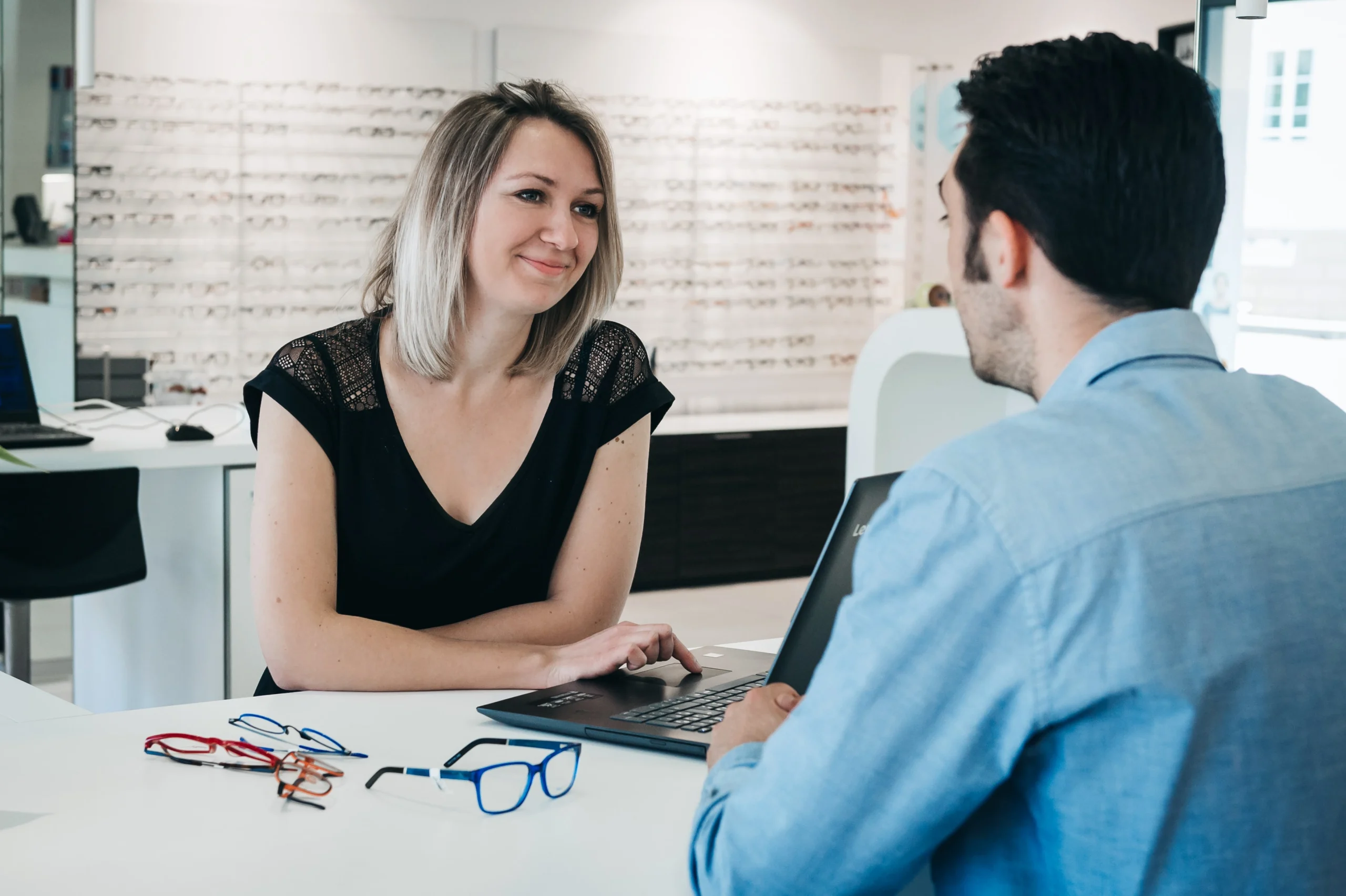 Une opticienne discute avec un client, ils sont assis autour d'un bureau sur lequel sont posées des lunettes de vue